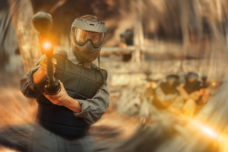 Woman in protective clothes and helmet aiming with paintball marker