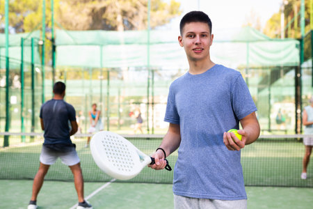 Positive sporty young guy standing on paddle tennis outdoor courtの写真素材