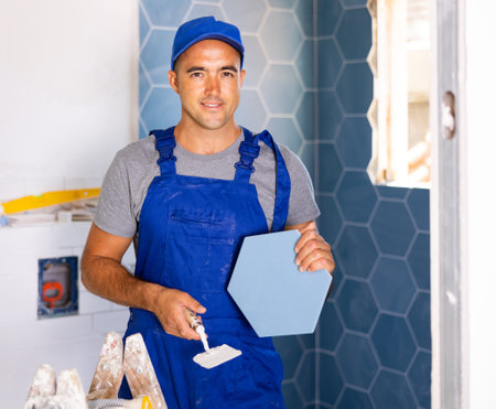 Portrait of young master wearing blue construction overalls posing with ceramic tiles and...