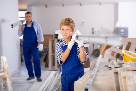 Boy in blue overalls carries an aluminum profile on his shoulder to renovate room