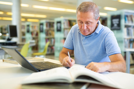 Focused casual mature male writing in notepad and reading books in the libraryの写真素材