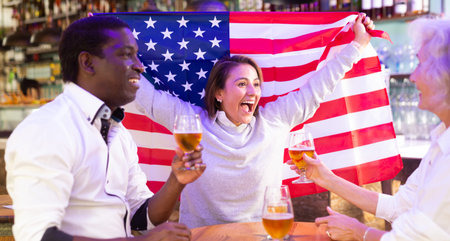 Happy international sport supporters holding up the flag of USA and drinking beer in the pubの写真素材