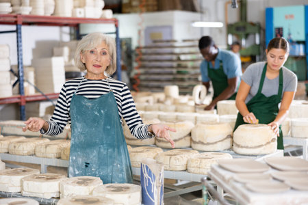 Portrait of an elderly craftswoman among ceramic plates and cups in the workshop of pottery factory