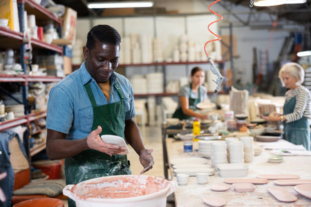 African american worker in pottery factory dips clay plates and cups into container with glazeの写真素材