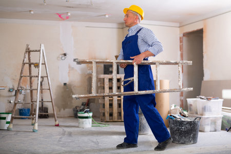 Worker in blue overalls carries a ladder on his shoulder in room being renovatedの写真素材