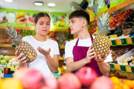 Male supermarket worker helping female consumer choosing pineapple in grocery shopの写真素材