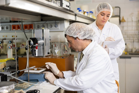Latin male scientist using microscope while female chemist taking notes in lab