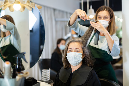 Woman hairdresser in protective mask cutting hair of elderly female clientの写真素材