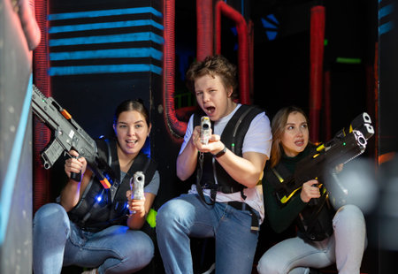 Two girls and one guy sitting and hiding behid cover while playing lasertagの写真素材
