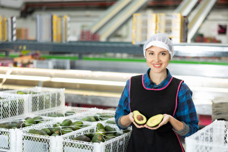 Smiling female worker in fruit sorting warehouse holding sliced avocado