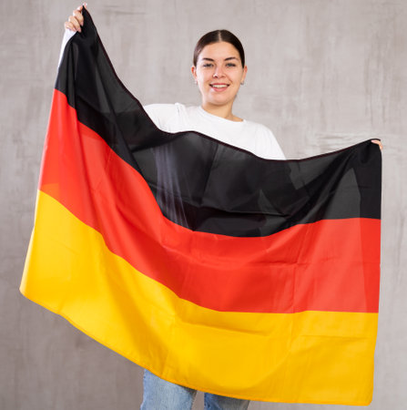 Cheerful patriotic young woman holding flag of Germany against gray background indoorsの写真素材
