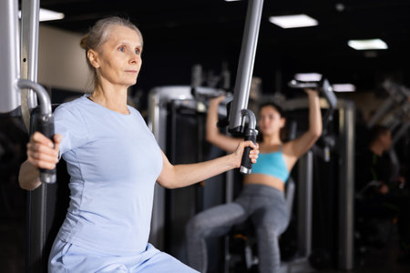 Elderly female training trains muscles of arms and press on simulator in gym
