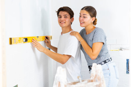 Positive couple checking laid tiles on wall with level meter in room under renovation