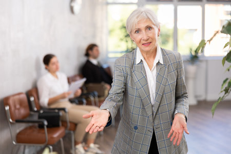 Smiling secretary senior woman stands at entrance to office reception and expects visitors
