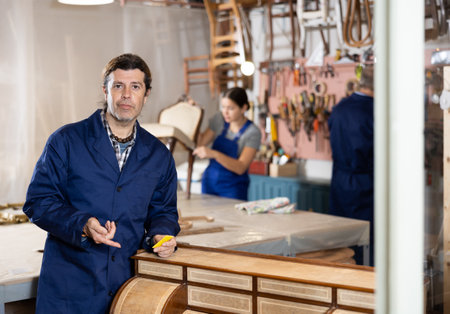 Professional middle-aged craftsman restorer in uniform renewing antique dresser while working in repair shopの写真素材