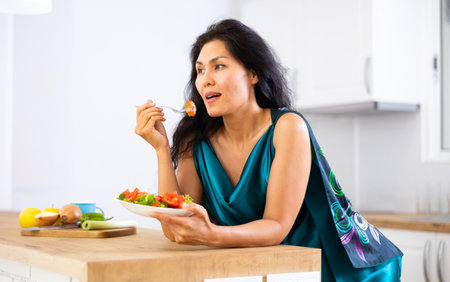 Woman leaning on kitchen table and eating saladの写真素材