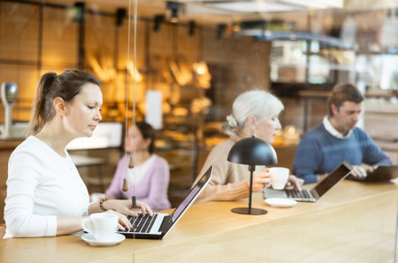 Focused woman drinking coffee while working on laptop in cozy cafeの写真素材
