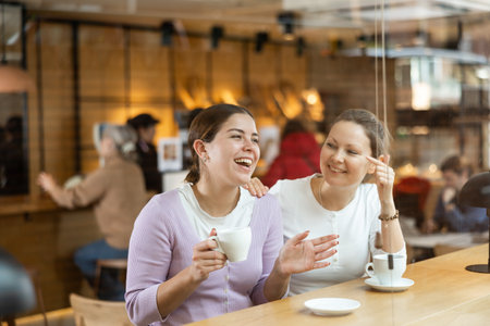 Positive young and middle-aged women in casual wear sitting at table and enjoying coffee or tea in cozy cafeteriaの写真素材
