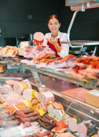 Smiling young girl seller offering fresh mortadella in grocery storeの写真素材