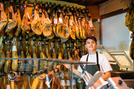 Young salesman in black apron offering dry-cured jamon in butcher shopの写真素材