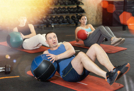 Strong middle-aged man performing exercise for abs core with medicine ball on yoga mat during group...