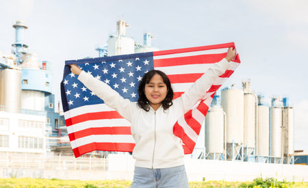 Happy young woman with flag of the USA against background of factory