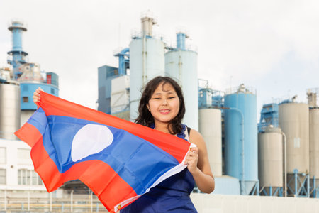 Young girl in blue overalls waving the flag of Laos against backdrop of modern metallurgical plantの写真素材