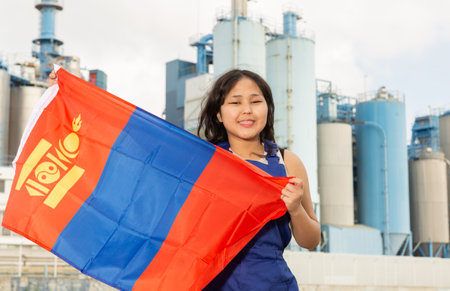 Portrait of positive girl in construction overalls with the Mongolia flag on the background of modern factoryの写真素材