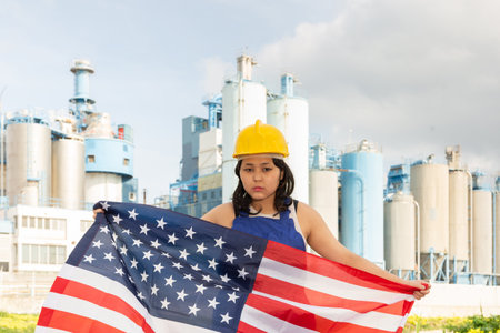 Worried asian girl in work clothes and hardhat with flag of usa standing in front of industrial sceneryの写真素材