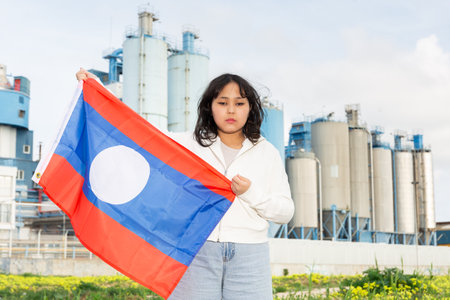 Sad young woman with flag of Laos against background of factoryの写真素材
