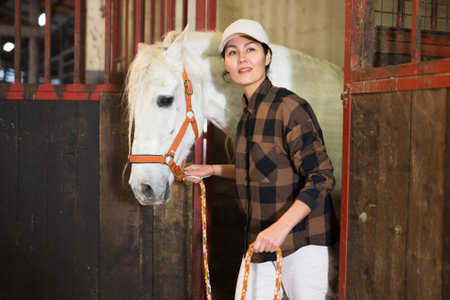 Positive woman leading white horse out of stall in a stable