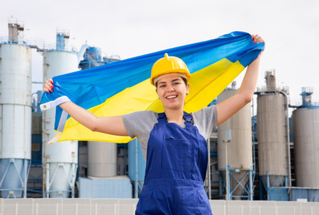 Young female engineer in helmet waving state flag of Ukraine while standing in front of big tanks at chemical plantの写真素材