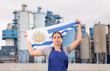 Sad young woman worker with flag of Uruguay against background of factoryの写真素材