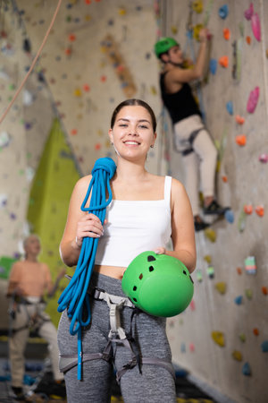 Smiling active young woman standing with rope on shoulders and helmet in hand against artificial training climbing wall in adventure parkの写真素材
