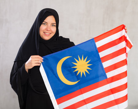 Balanced smiling Muslim woman in traditional black hijab holds flag of Malaysiaの写真素材