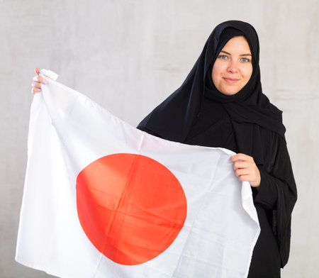 Balanced Muslim woman in traditional black hijab holds flag of Japanの写真素材