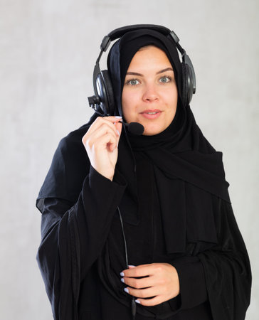 smiling Muslim woman in black hijab and headphones with microphone on gray background.