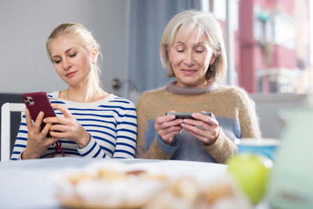 Mature mother and daughter together playing on their smartphones while sitting at table at homeの写真素材