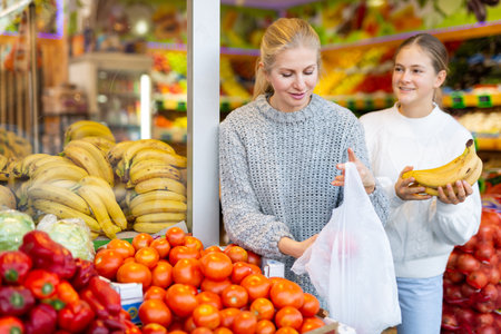 Mom picks tomatoes while daughter buys bananas at grocery storeの写真素材