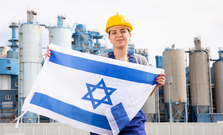 Young female engineer in helmet waving state flag of Israel while standing in front of big tanks at chemical plantの写真素材