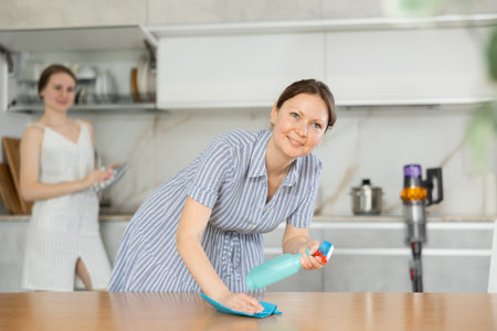 Pleased mother dusting table while daughter washes dishes in kitchen