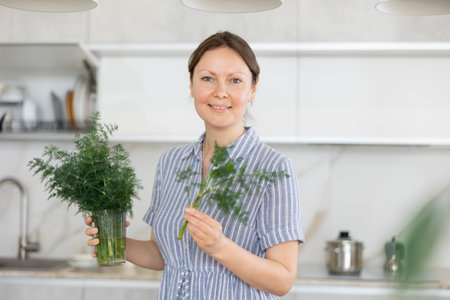 Woman in dress with short sleeves sitting near kitchen table and holding glass with bunch of dillの写真素材