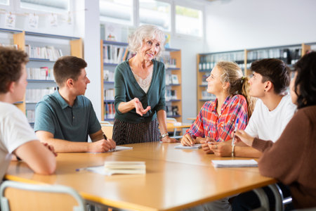 Mature female teacher working with friendly group of schoolgirls and schoolboys studying in libraryの写真素材