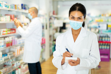 Portrait of an female pharmacist in protective mask, working in pharmacy during the pandemic; standing in trading floor and makes important notesの写真素材