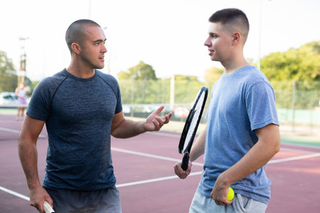 Two men with rackets in their hands chatting after playing padel on tennis court outdoorの写真素材
