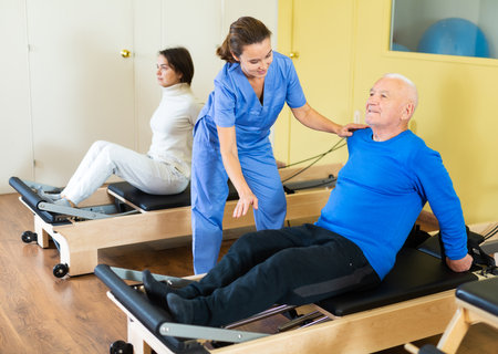 Medical professional helps and monitors the difficulty of Pilates exercise for an mature manの写真素材