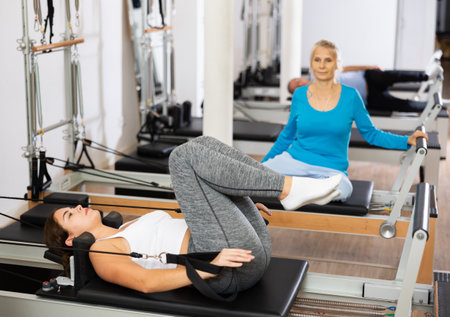 Young European female training lower back while laying and pulling with legs bands of Pilates reformer machine bed at gym. Pilates and healthy lifestyle conceptの写真素材