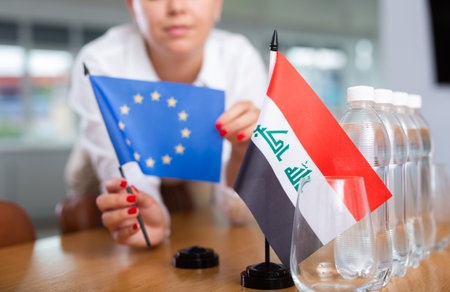 Woman secretary prepares an office for negotiations - she places flags of Iraq and European Union tableの写真素材