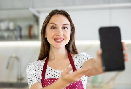 At home in kitchen smiling woman in apron shows blank smartphone screen close up. Dark touch screen workspaceの写真素材