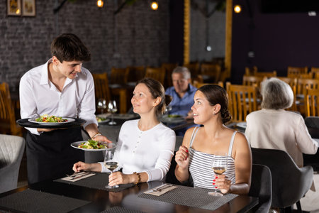 Young male waiter serving dish to two women in restaurantの写真素材